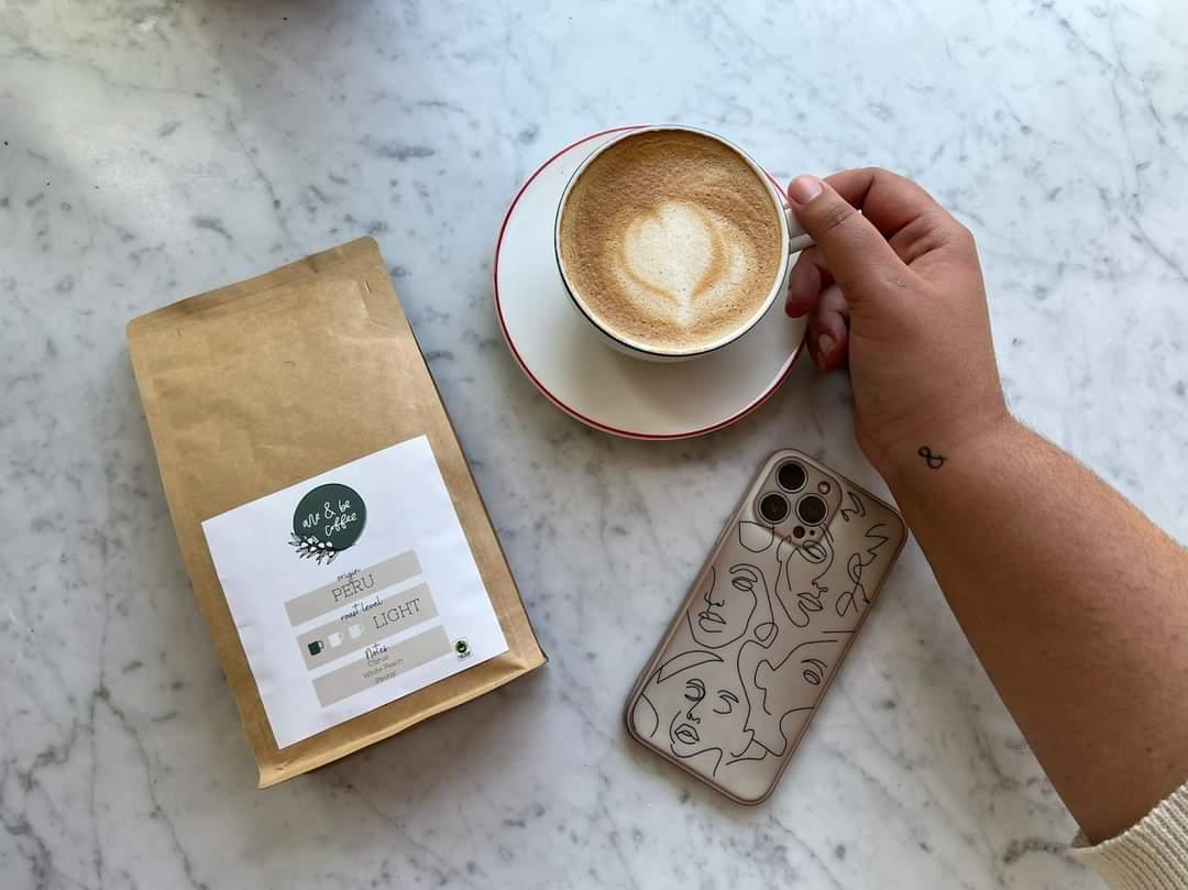 an iphone, a bag of are and be coffee, and a hand holding a round coffee cup and saucer on a marble surface