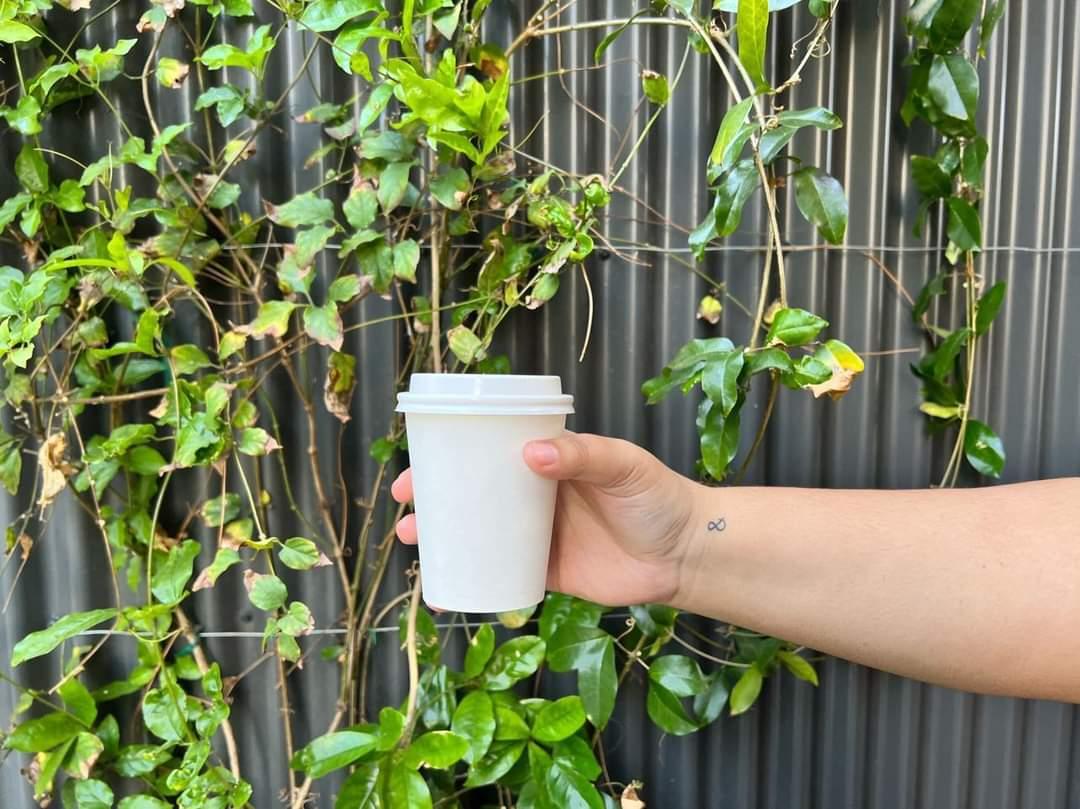 a hand holding a white takeaway cup in front of green plants and a metallic grey background